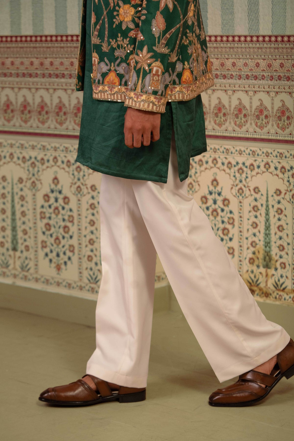 A person wearing a dark green embroidered traditional bomber with gold detailing, paired with white trousers and brown leather loafers, standing in front of a patterned wall.