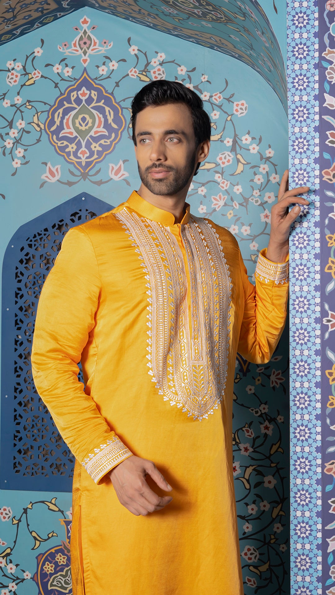 Man wearing a mustard traditional outfit with intricate patterns against a decorative blue wall.