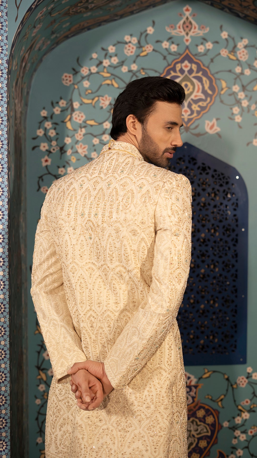 Man wearing a cream sherwani in front of an ornate wall.