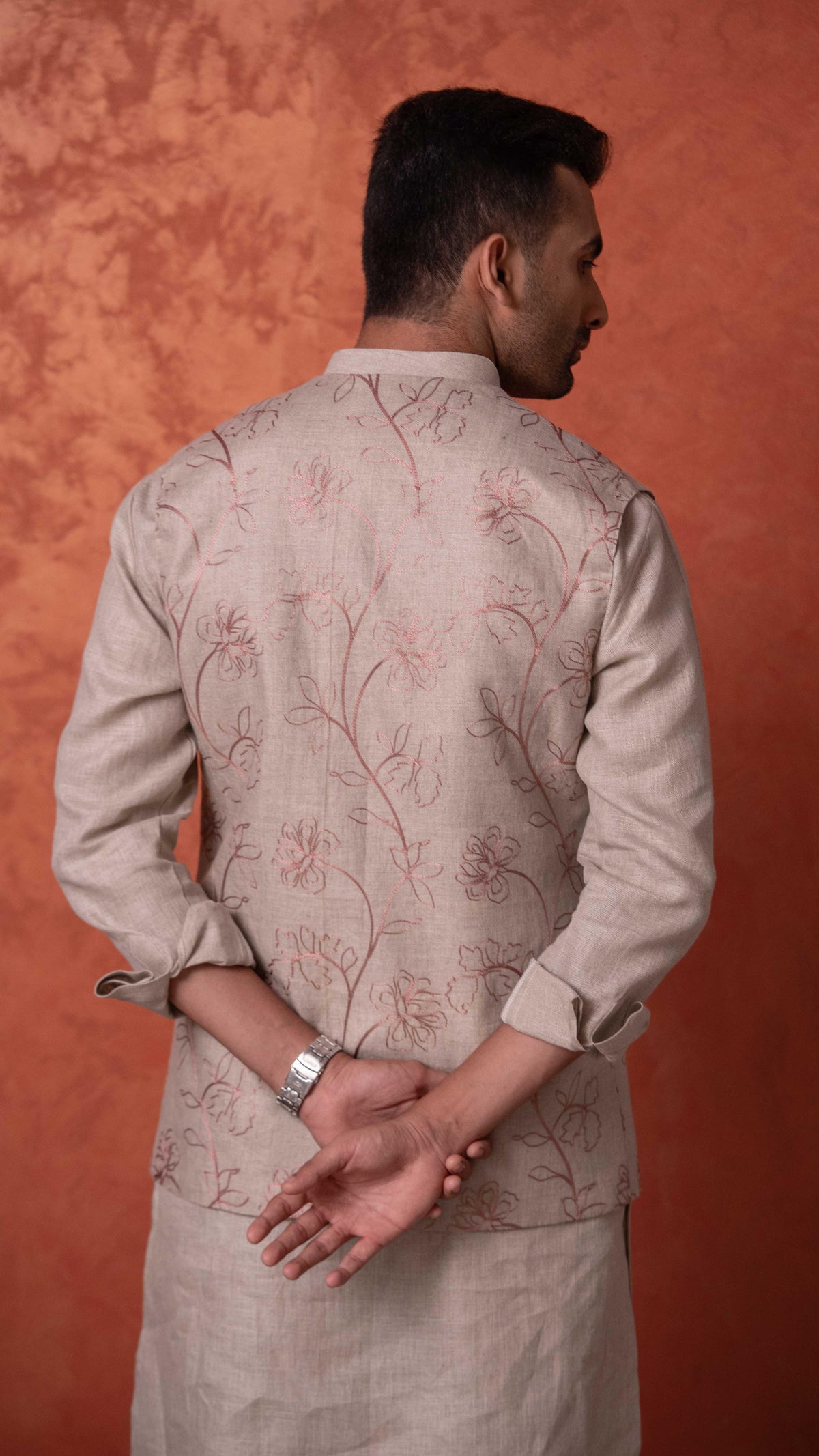 A man shown from the back wearing a light beige kurta with delicate brown floral embroidery, set against a textured orange-brown studio backdrop.