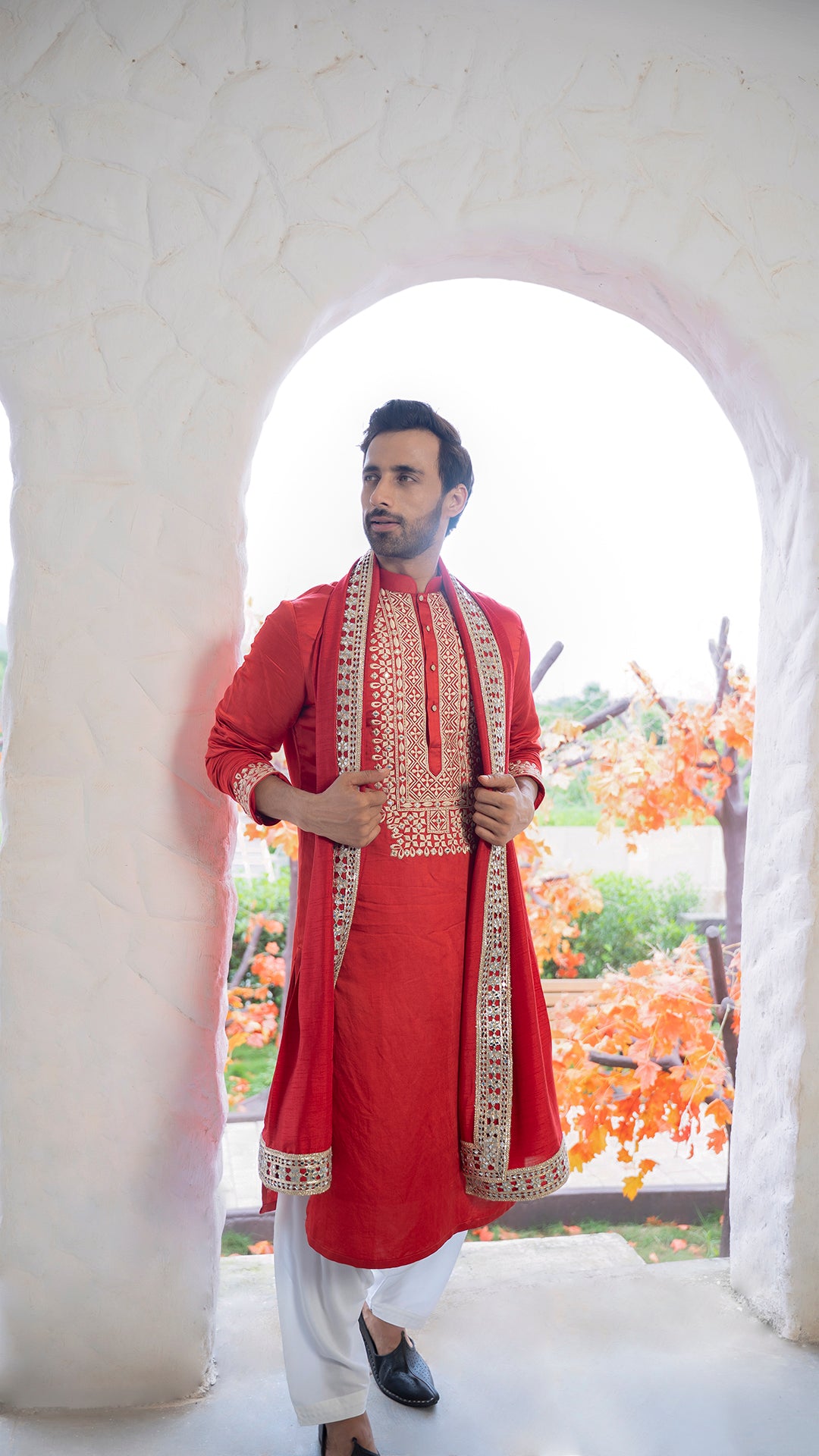 Man wearing a red kurta with beige embroidery standing in a doorway.