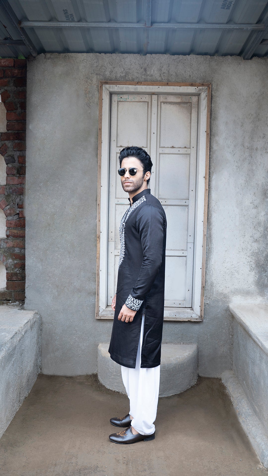 Man in a black kurta with white patiala standing in a room with a textured wall and window.