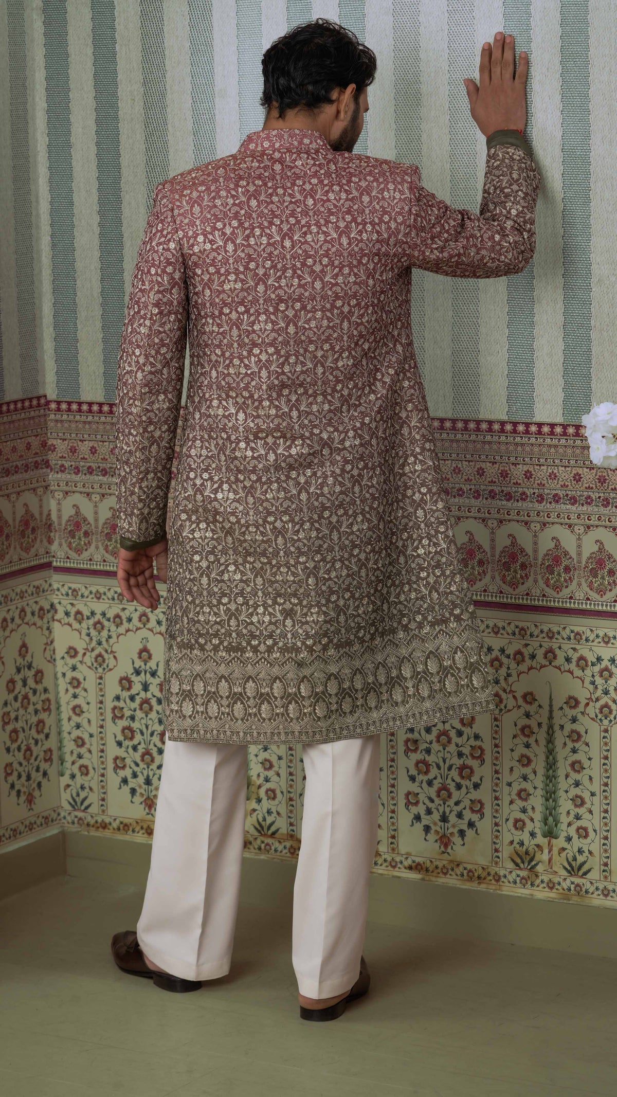 A man viewed from behind wearing a long-sleeved, heavily embroidered sherwani in a orange green and gold pattern, paired with off-white trousers, standing against a patterned wallpaper background.