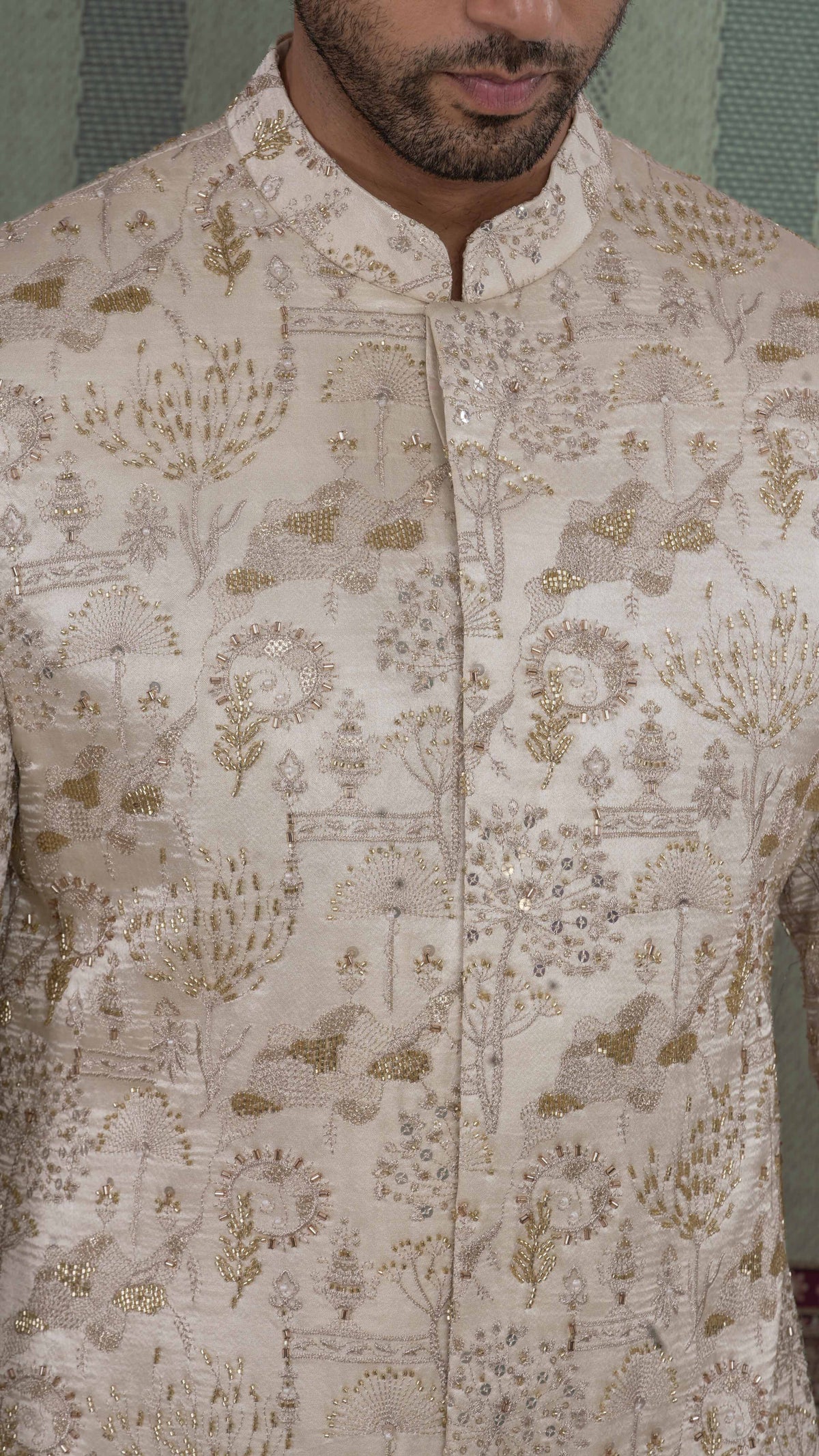 A close-up of a man wearing a beige-colored sherwani jacket with intricate gold and silver embroidery and mirror work, featuring a mandarin collar and buttoned front.