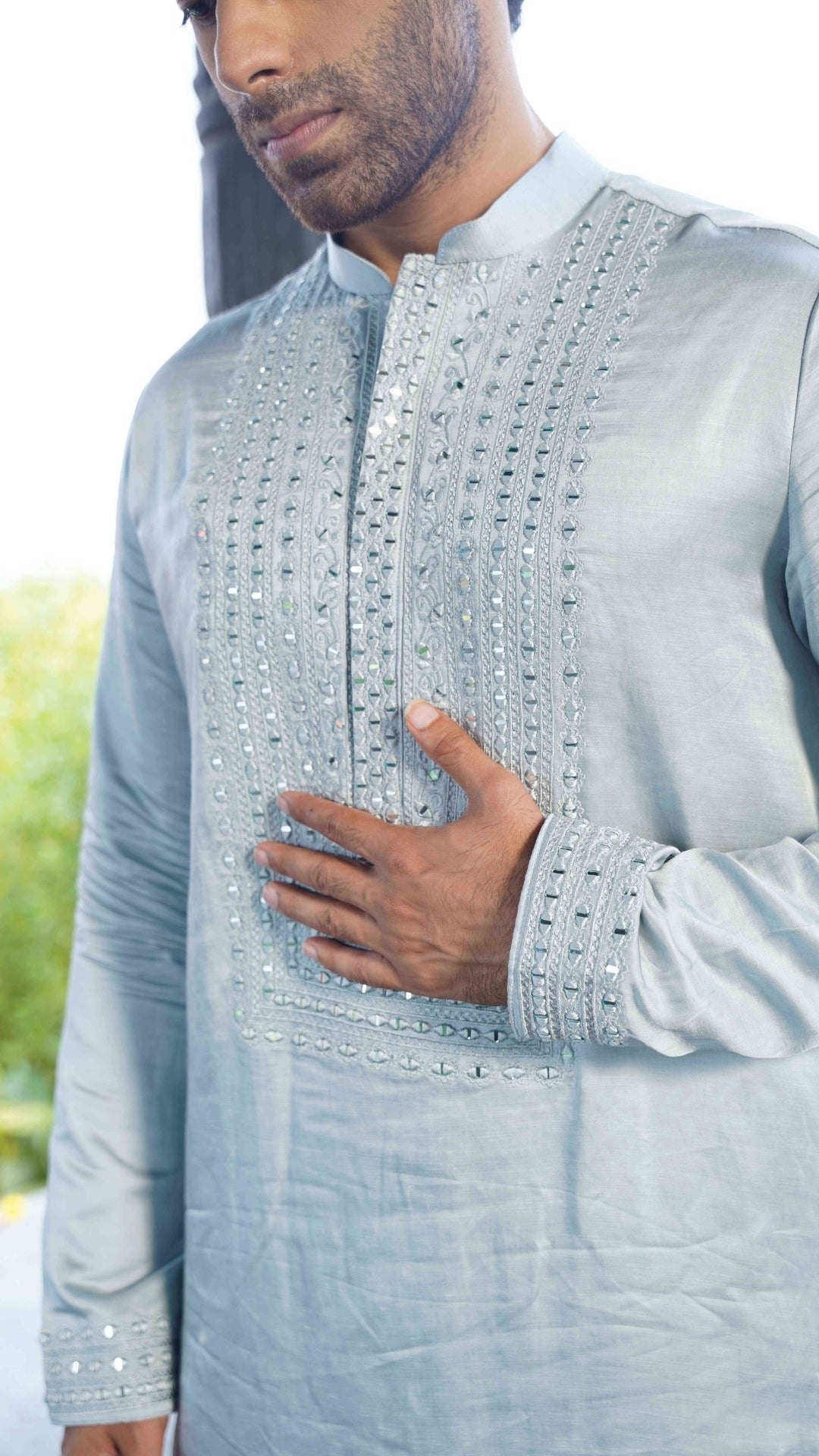 Man wearing a light blue embroidered traditional outfit with blurred greenery in the background
