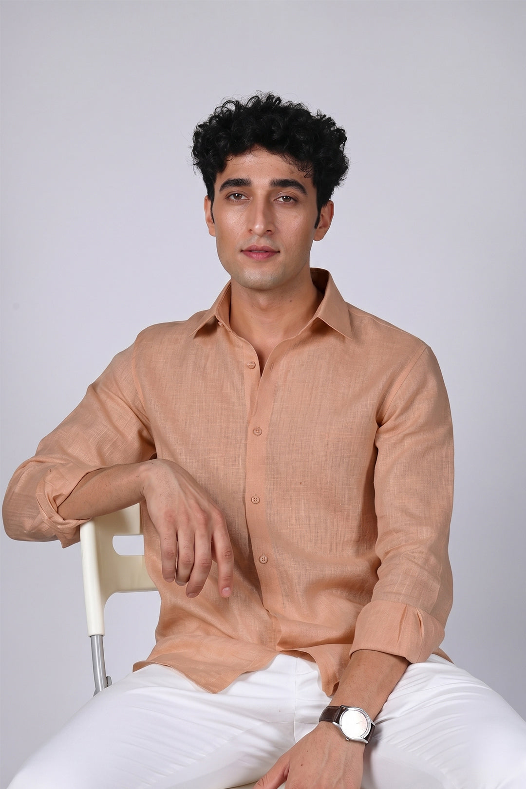 Peach Sandstone pure linen shirt for men, seated relaxed pose with arm resting on chair, spread collar and long sleeves, styled with white trousers, by BWO