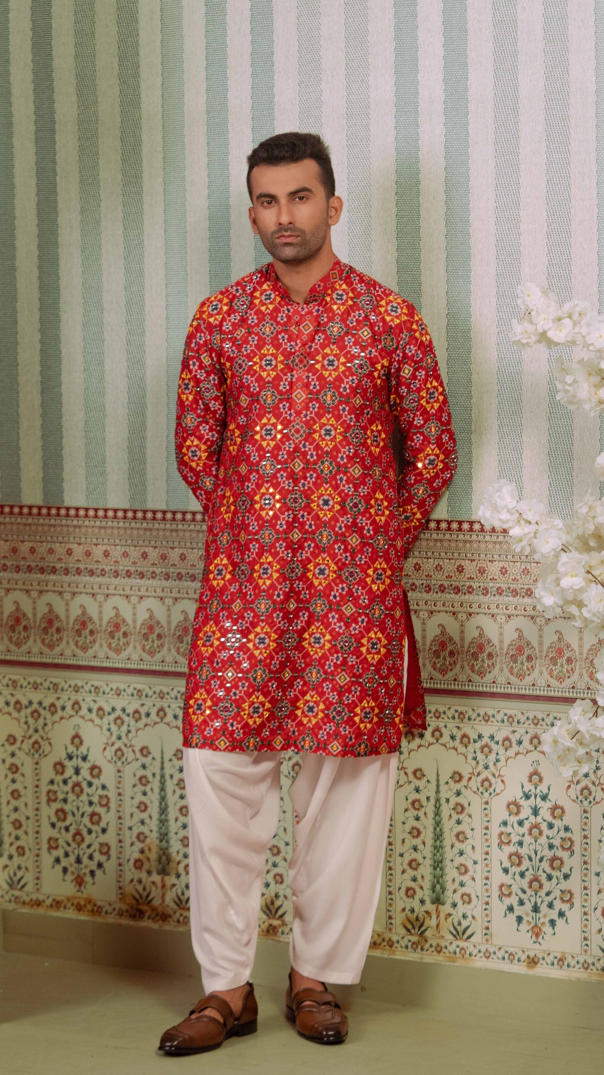 A man wearing a red printed patola kurta with yellow, orange, and blue patterns, paired with off-white trousers and brown leather loafers, standing against a patterned green and white wall.