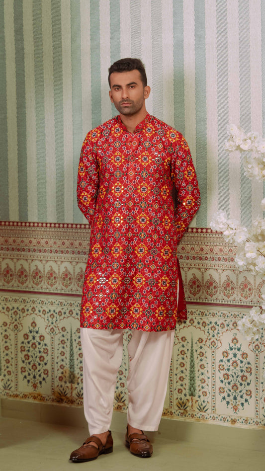 A man wearing a red printed patola kurta with yellow, orange, and blue patterns, paired with off-white trousers and brown leather loafers, standing against a patterned green and white wall.