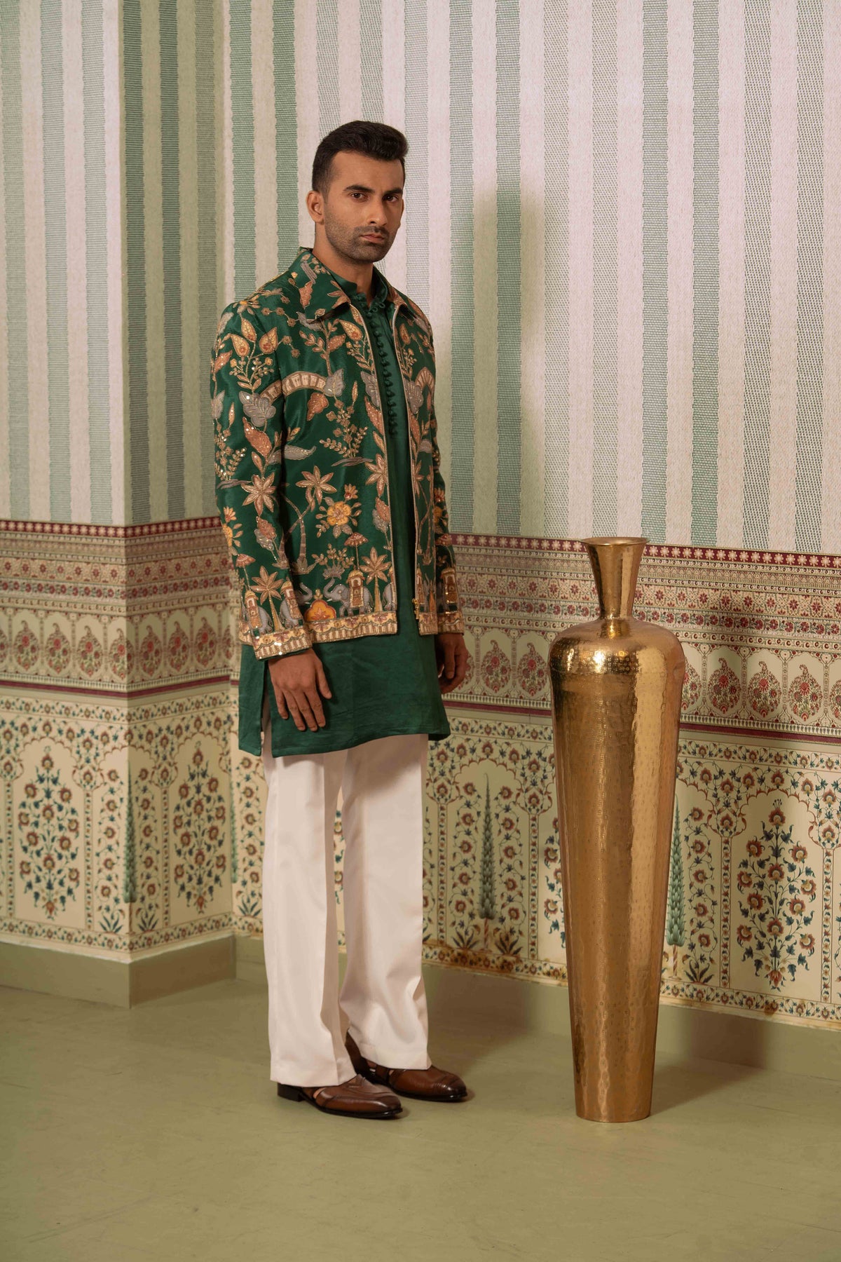 A man modeling a traditional South Asian outfit featuring a green bomber jacket with intricate gold floral embroidery, paired with white trousers and brown leather shoes, standing next to a large gold decorative vase against a patterned wallpaper background.