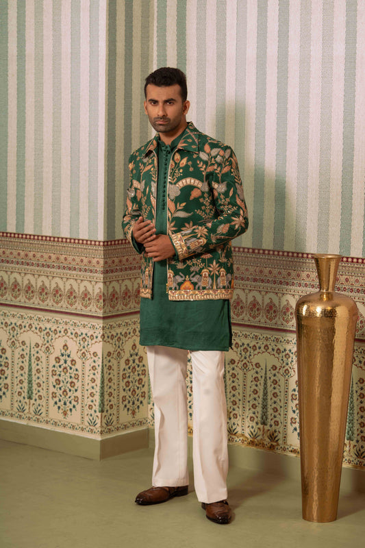 A man modeling a traditional South Asian outfit featuring a green bomber floral printed jacket, a solid green kurta, off-white trousers, and brown shoes, standing in front of a patterned wallpaper background with a large gold vase.