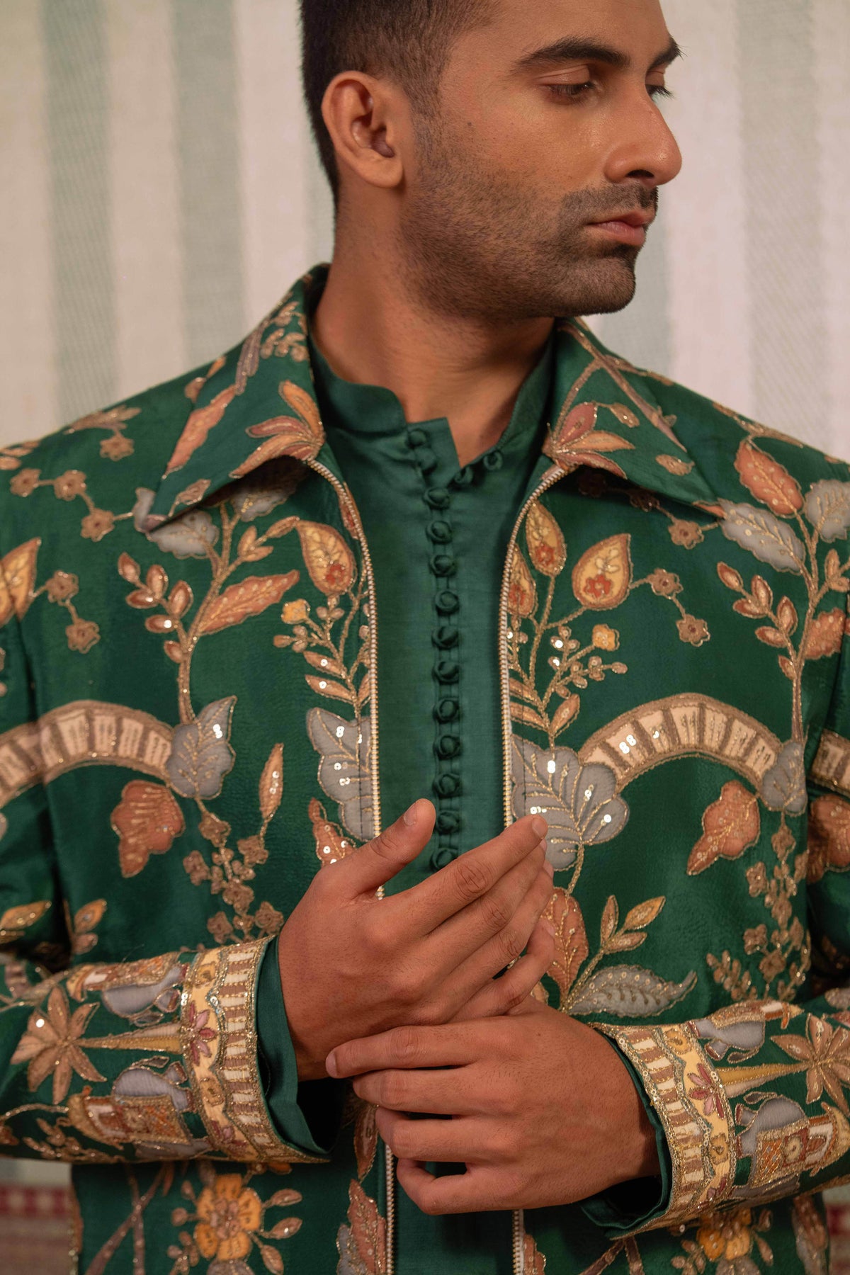 A man wearing a dark green embroidered traditional jacket with intricate gold, beige, and brown floral patterns over a matching green shirt.