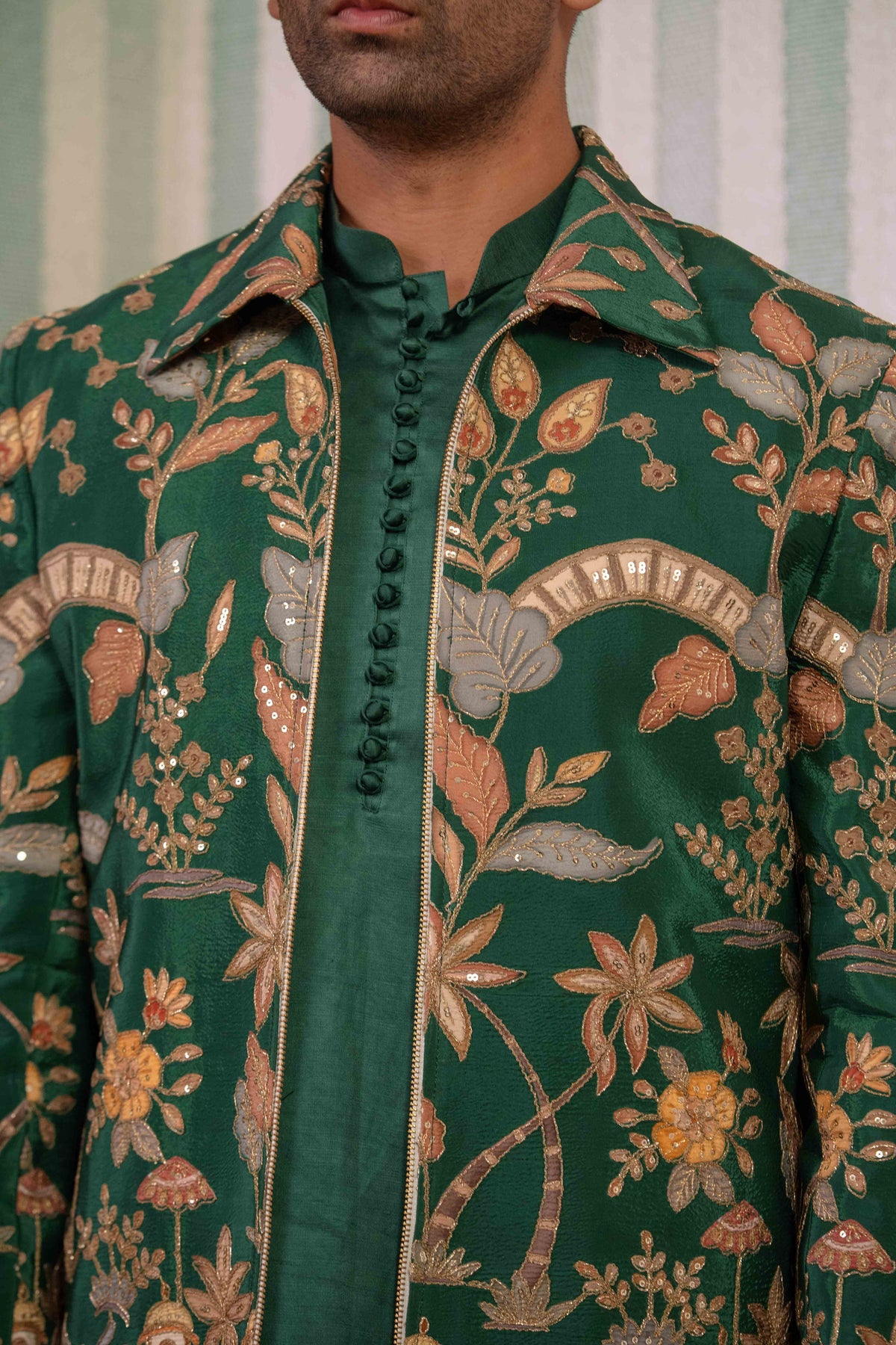 A close-up of a man wearing a dark green jacket with an intricate floral and botanical pattern in gold, beige, and brown tones, paired with a matching green shirt.