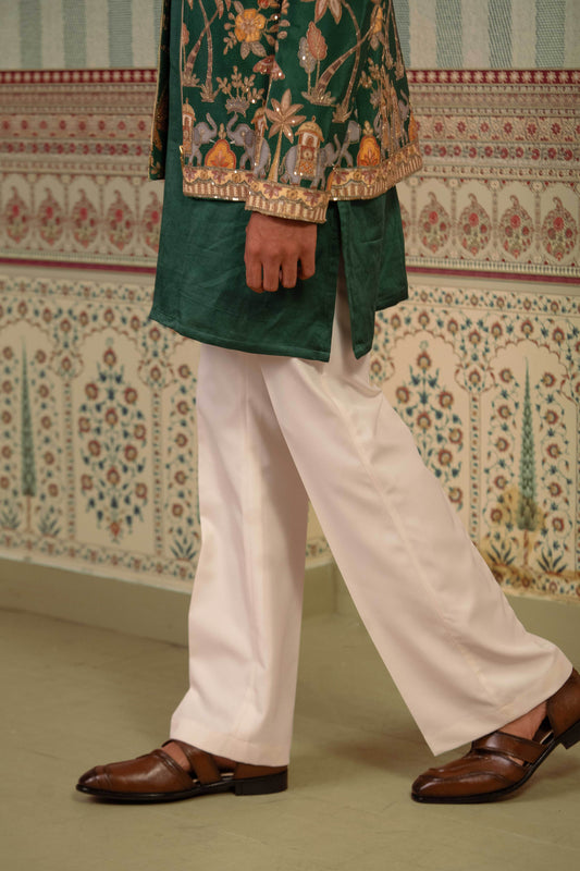 A person wearing a dark green embroidered traditional bomber with gold detailing, paired with white trousers and brown leather loafers, standing in front of a patterned wall.