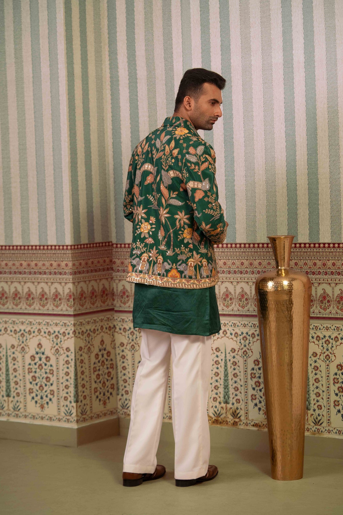 A man wearing a green floral printed kurta with a solid green waist sash and white trousers, standing in front of a patterned wallpaper background next to a large gold vase.