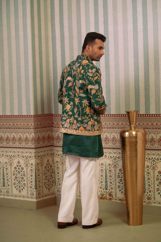 A man wearing a green floral printed kurta with a solid green waist sash and white trousers, standing in front of a patterned wallpaper background next to a large gold vase.
