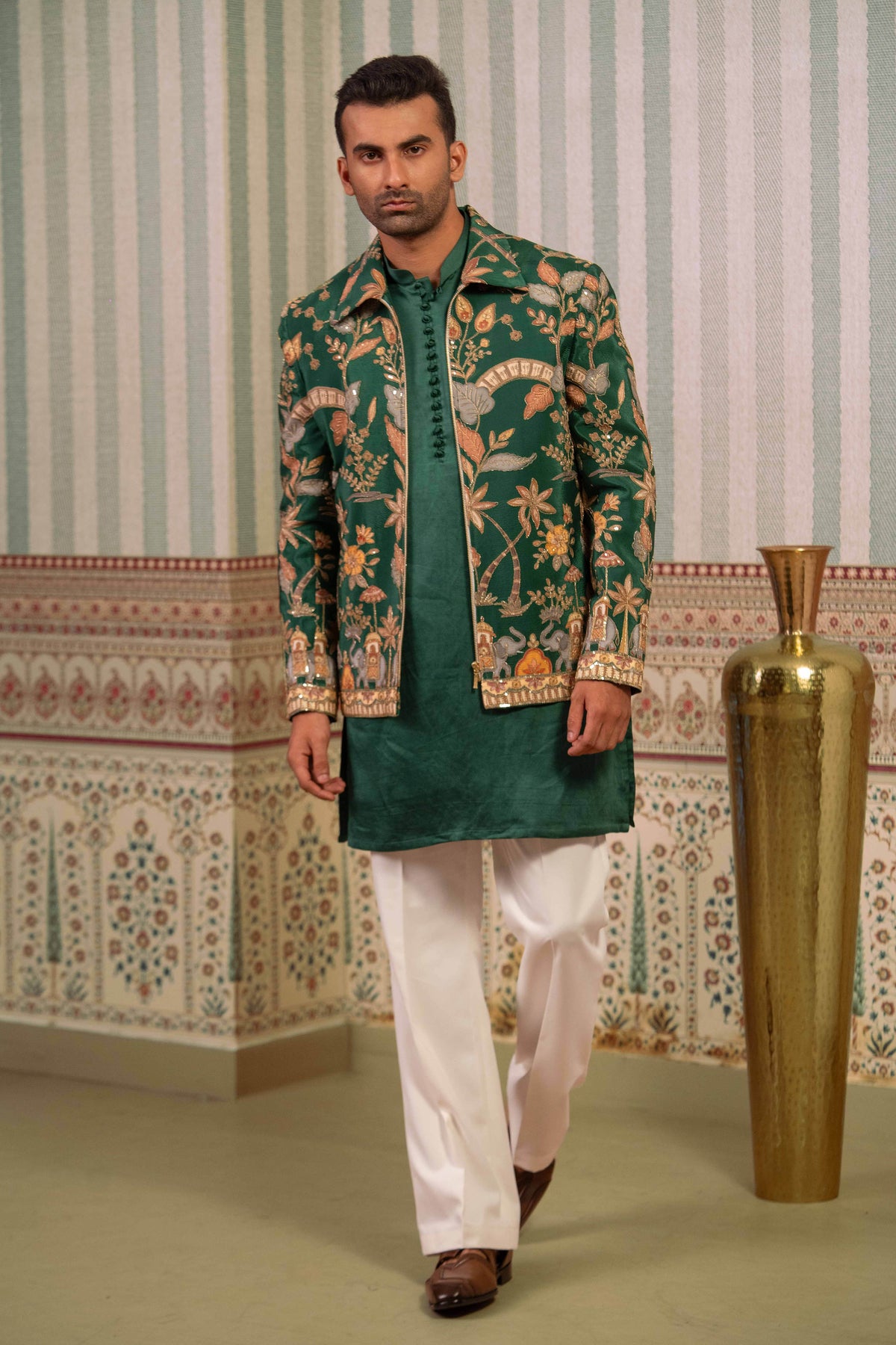 A man modeling a traditional Indian outfit featuring a green bomber, a green and gold patterned jacket, white trousers, and brown shoes, set against a patterned wall with a large gold vase.