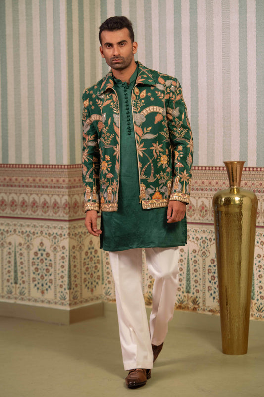 A man modeling a traditional Indian outfit featuring a green bomber, a green and gold patterned jacket, white trousers, and brown shoes, set against a patterned wall with a large gold vase.