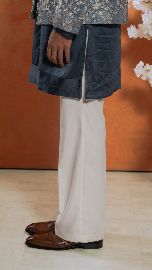 A person wearing a dark blue patterned tunic, off-white trousers, and brown leather shoes, standing against a warm orange-brown studio backdrop with white floral decor on the right.
