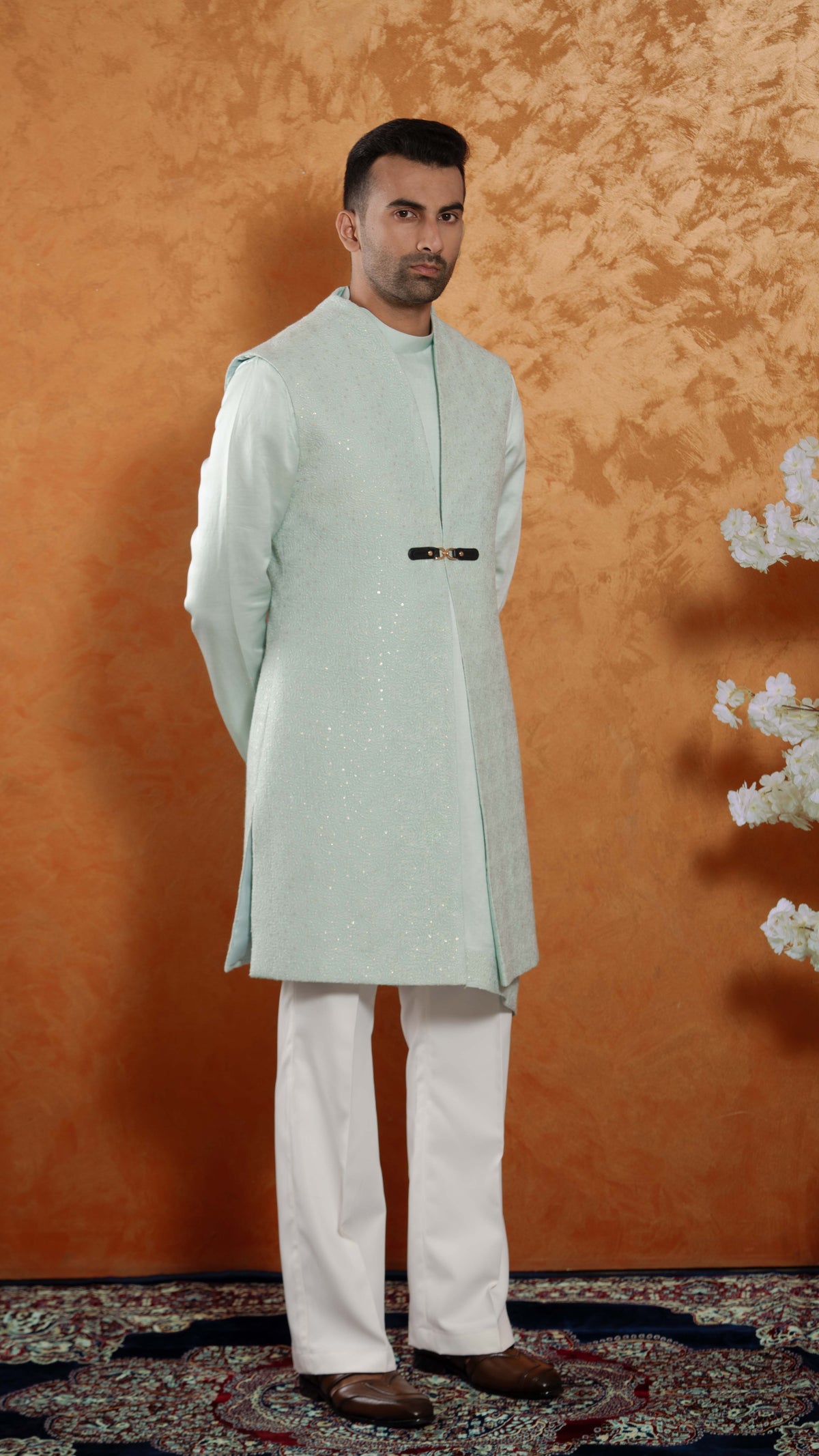 A man modeling a traditional light mint green sherwani with white trousers, standing against a textured orange-brown backdrop.