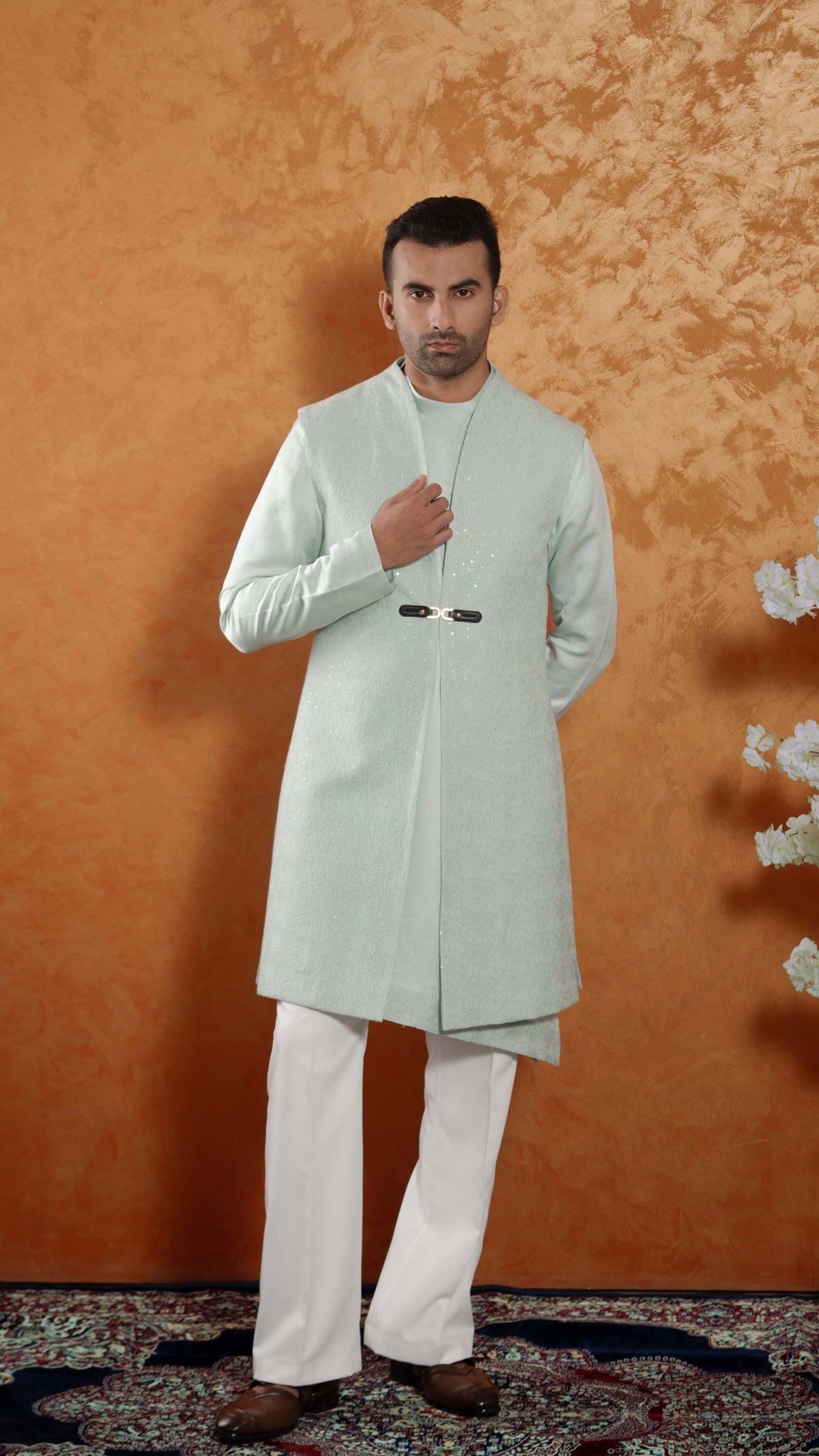 A man modeling a traditional light mint green bundi with white trousers, standing against a textured orange-brown backdrop.