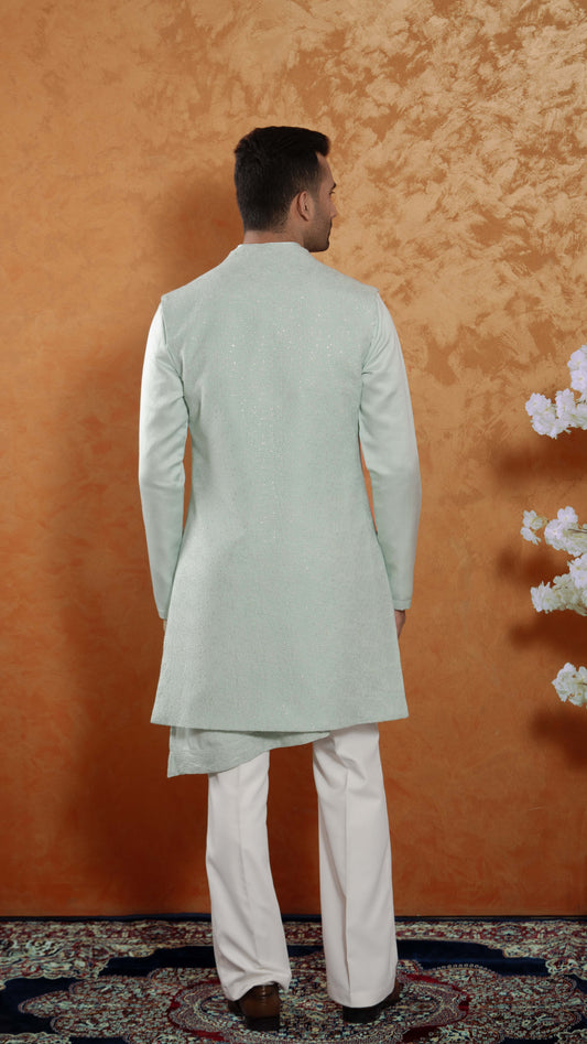 A man viewed from the back wearing a light mint green bundi with white trousers, standing against a textured orange-brown wall.