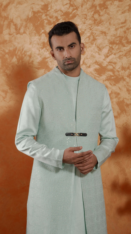 A man wearing a light mint green traditional long kurta with a black waist buckle, standing against a textured orange-brown studio backdrop.