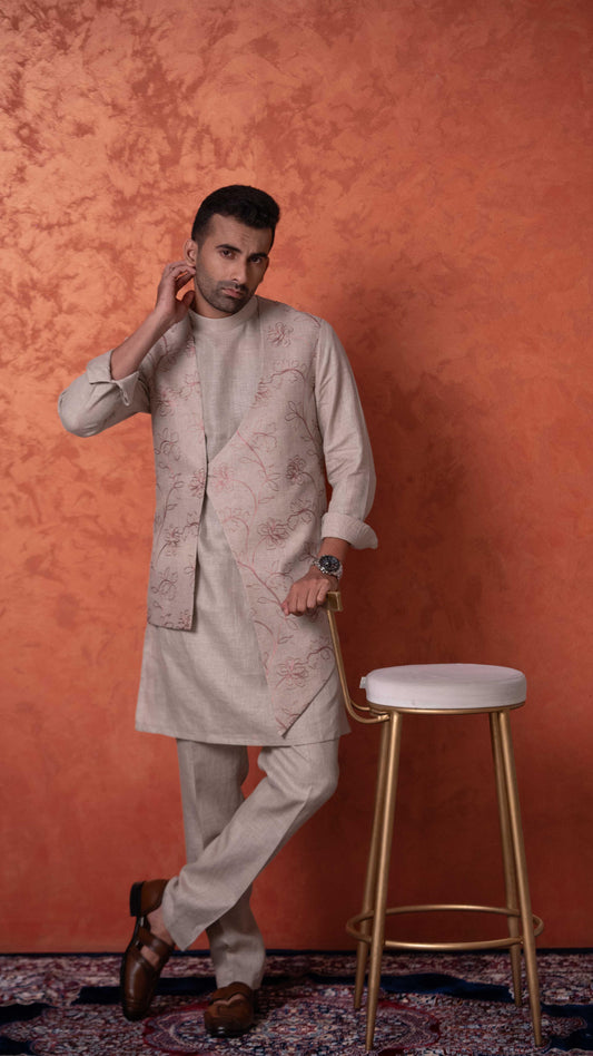 A man wearing a light beige printed kurta and matching trousers stands in a studio setting against a textured orange wall, leaning on a gold-framed stool.