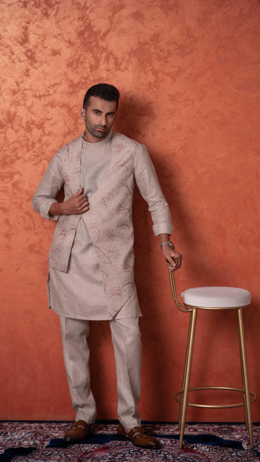 A man wearing a light beige traditional kurta and matching trousers stands against a textured orange wall, holding the arm of a gold-framed stool.
