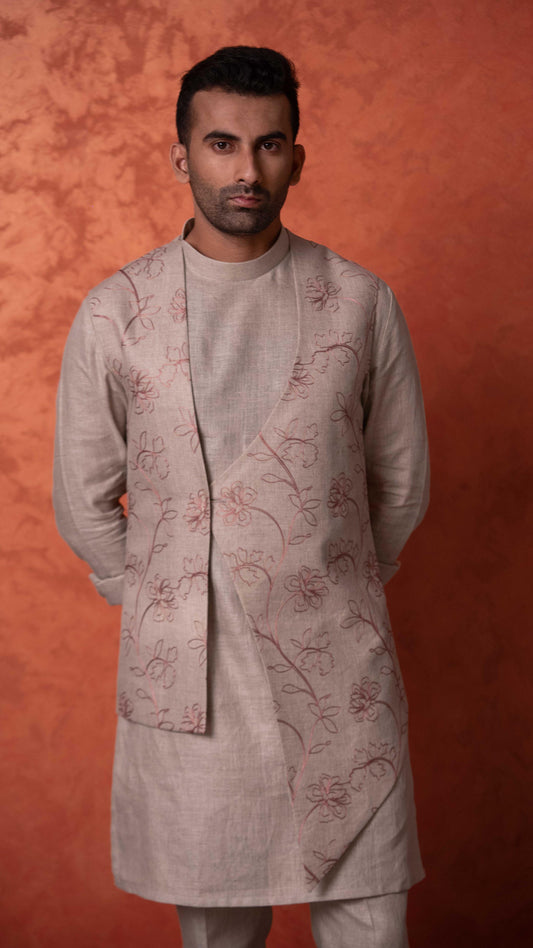 A man wearing a light beige kurta with brown floral embroidery, standing against a textured orange-brown studio backdrop.