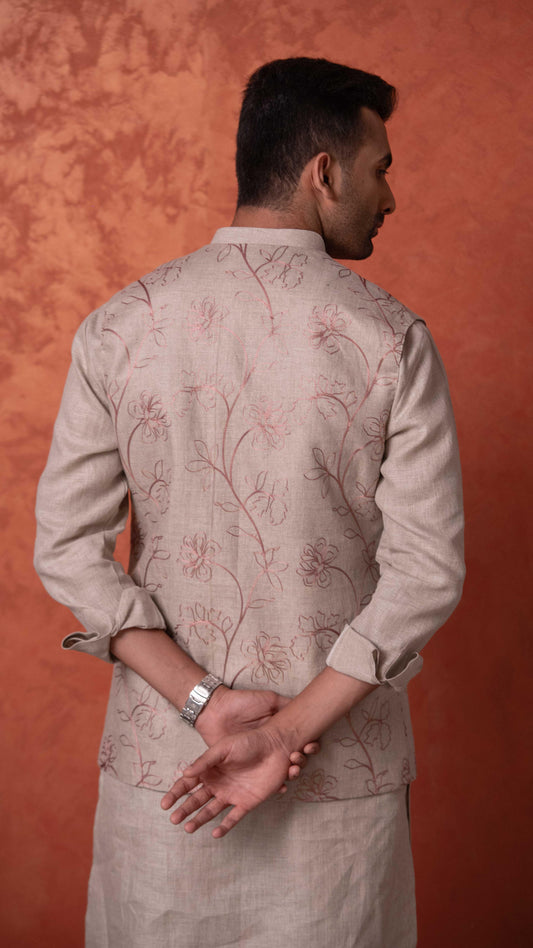 A man shown from the back wearing a light beige kurta with delicate brown floral embroidery, set against a textured orange-brown studio backdrop.