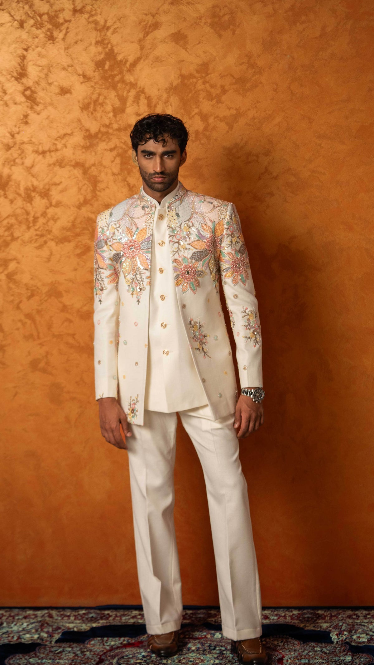 A man wearing a cream-colored traditional Indian sherwani jacket and matching trousers, featuring intricate multicolor floral embroidery on the chest and shoulders, standing against a textured orange-brown backdrop.