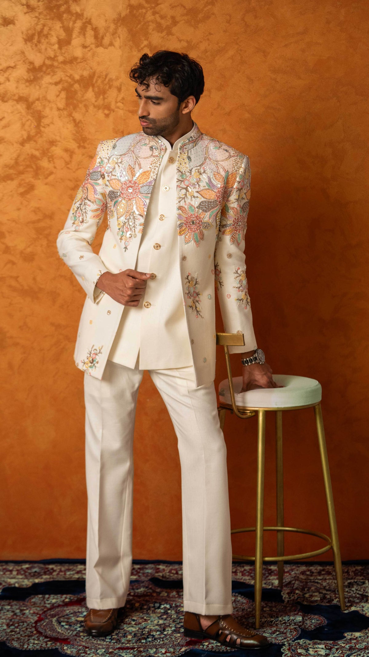 A man models a cream-colored traditional Indian suit with intricate multicolor floral embroidery on the jacket, paired with matching trousers, standing next to a gold-framed stool against a textured orange background.