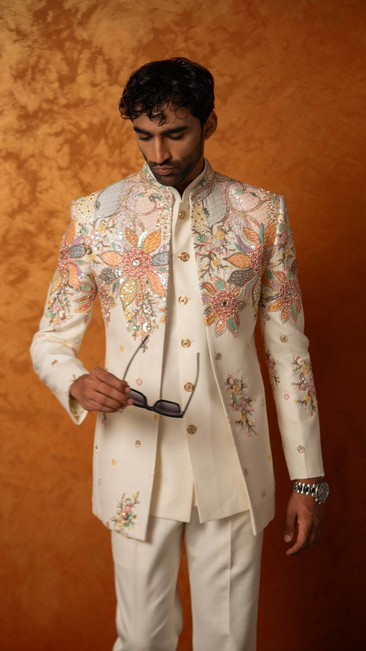 A man wearing a cream-colored traditional Indian sherwani jacket and matching trousers, decorated with intricate multicolor floral embroidery. He is holding a pair of sunglasses against a textured orange-brown studio backdrop.