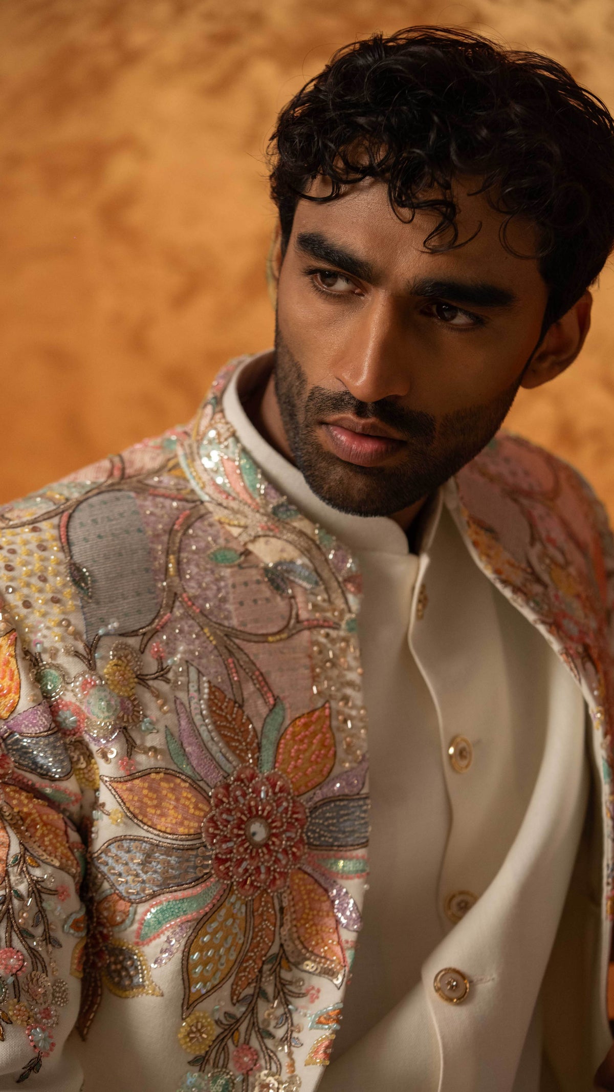 A close-up portrait of a man wearing a cream-colored traditional jacket with intricate multicolor floral embroidery and sequins, set against a warm, textured golden-brown background.