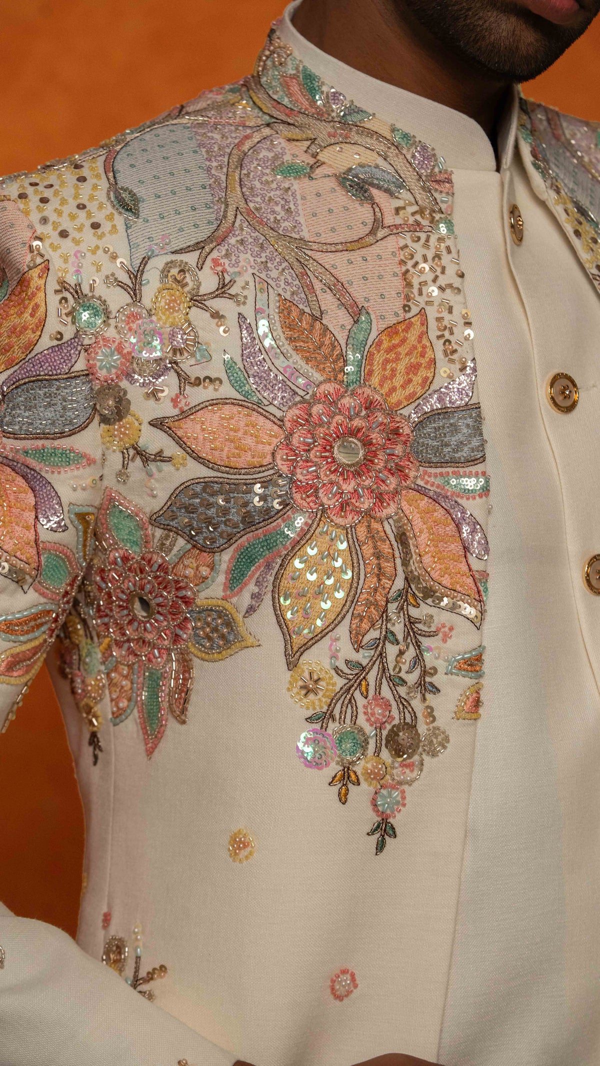 Close-up of a cream-colored traditional sherwani jacket with intricate multicolor floral embroidery and sequins on the chest, worn by a model against an orange background.