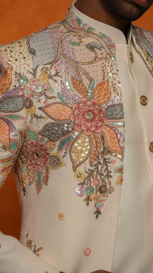 Close-up of a cream-colored traditional sherwani jacket with intricate multicolor floral embroidery and sequins on the chest, worn by a model against an orange background.