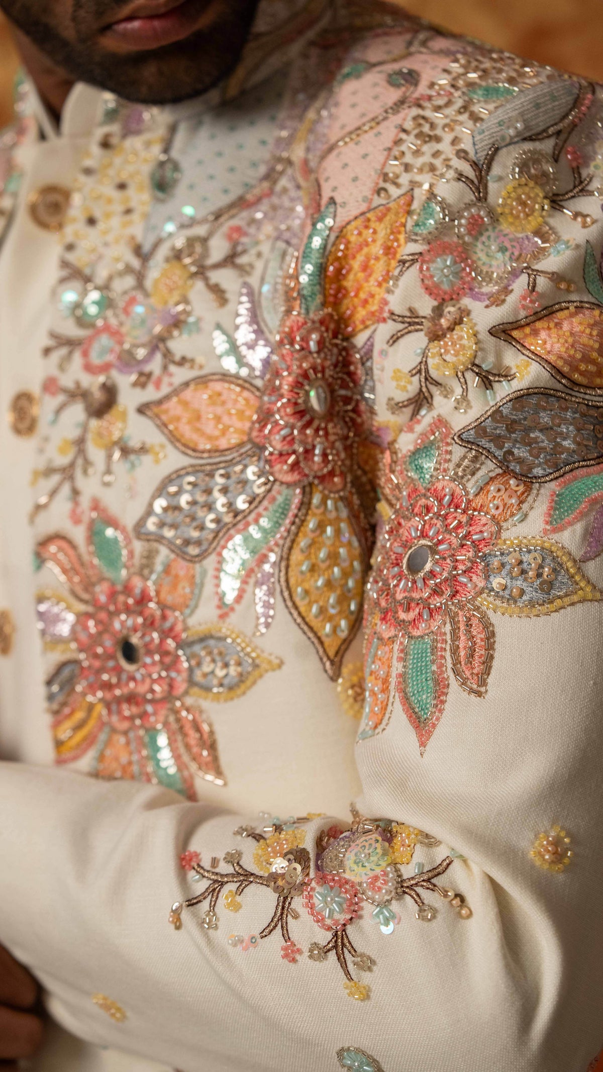 Close-up of a cream-colored traditional embroidered garment with intricate multicolor floral embroidery, sequins, and beadwork on the chest and sleeve.