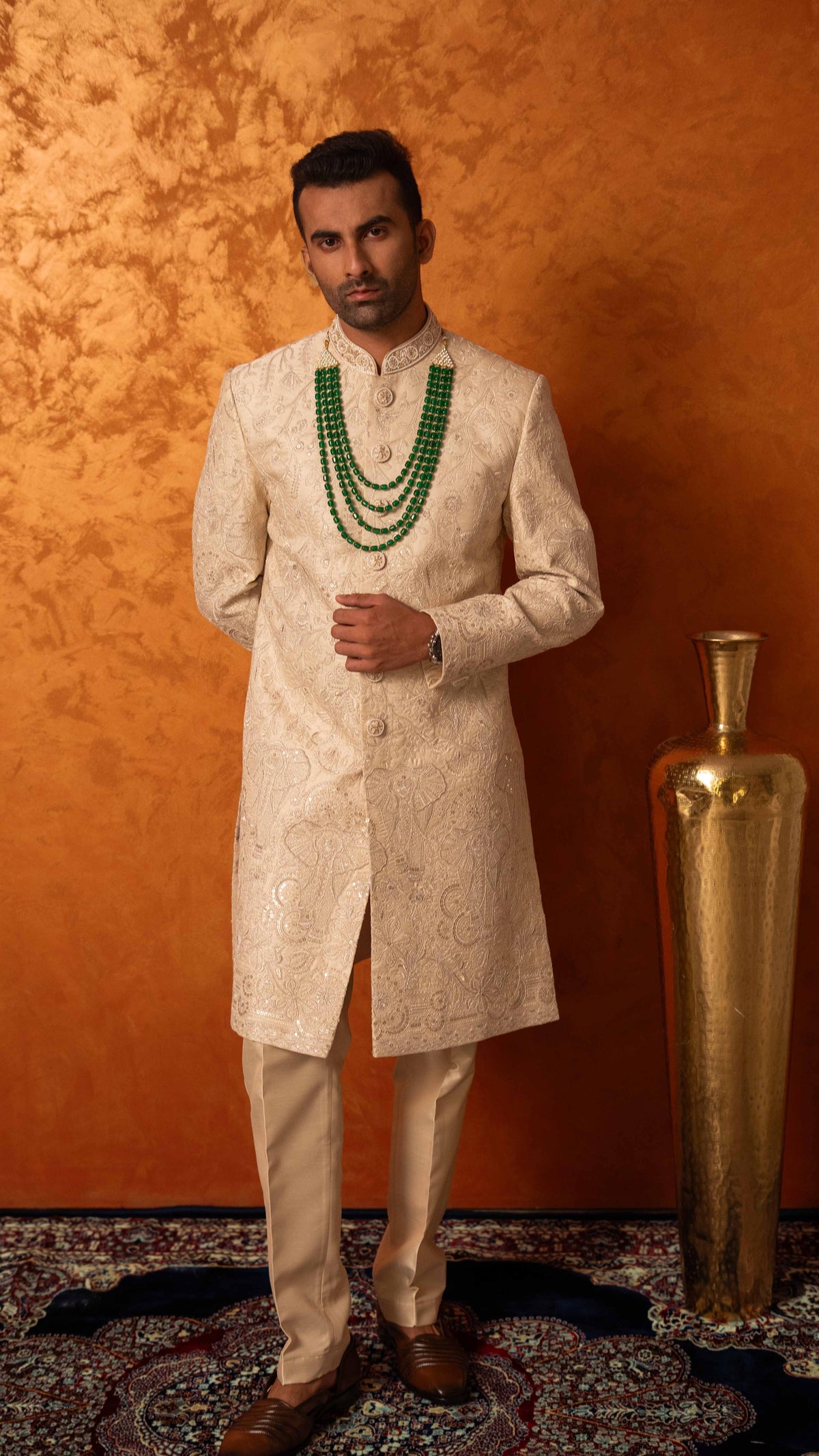 A man wearing a cream-colored embroidered sherwani and matching trousers, accessorized with green beaded necklaces, standing against a textured orange wall next to a gold decorative vase.