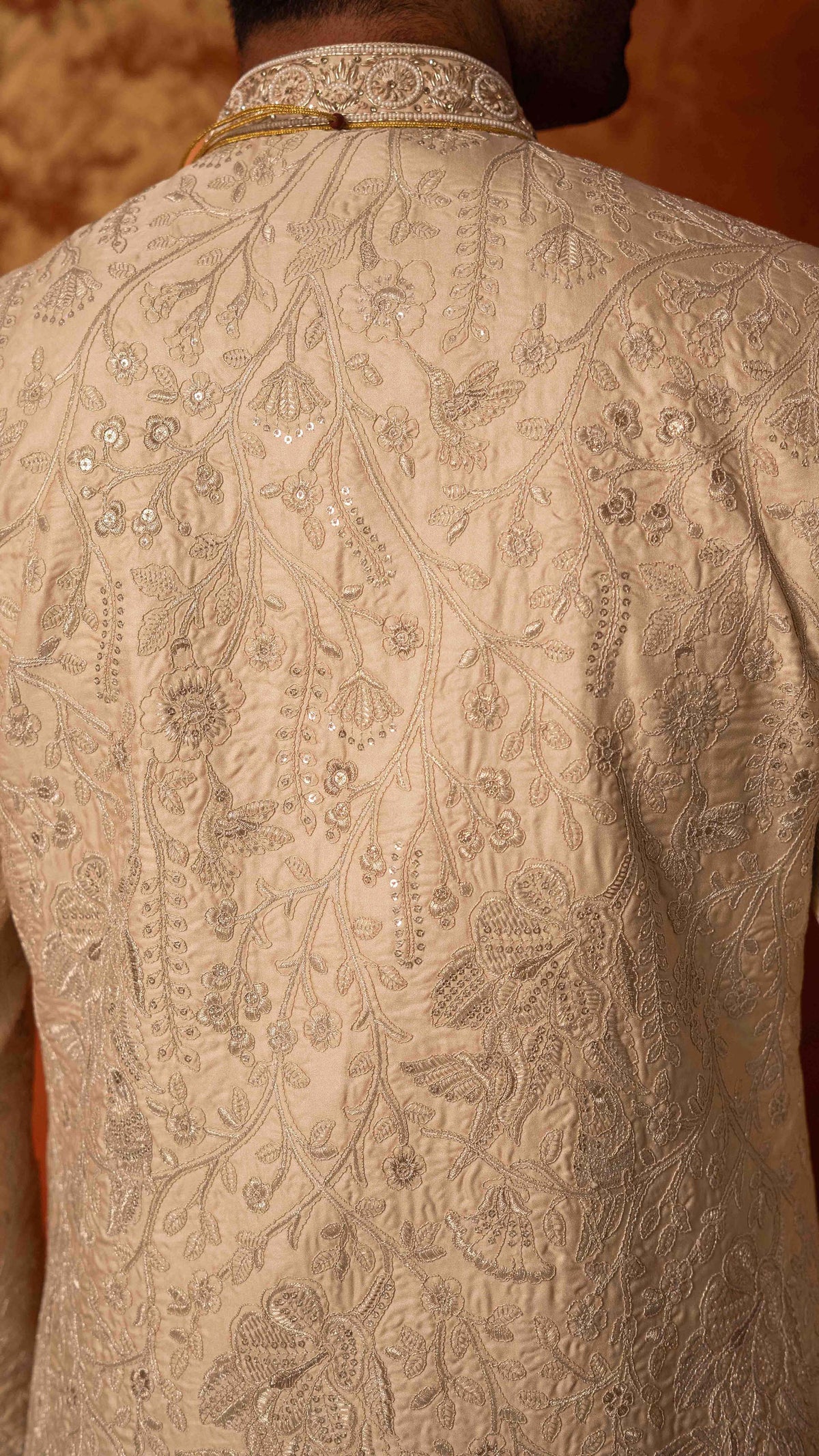 Close-up back view of a person wearing a cream or beige heavily embroidered sherwani jacket with intricate floral and vine patterns and sequined detailing.