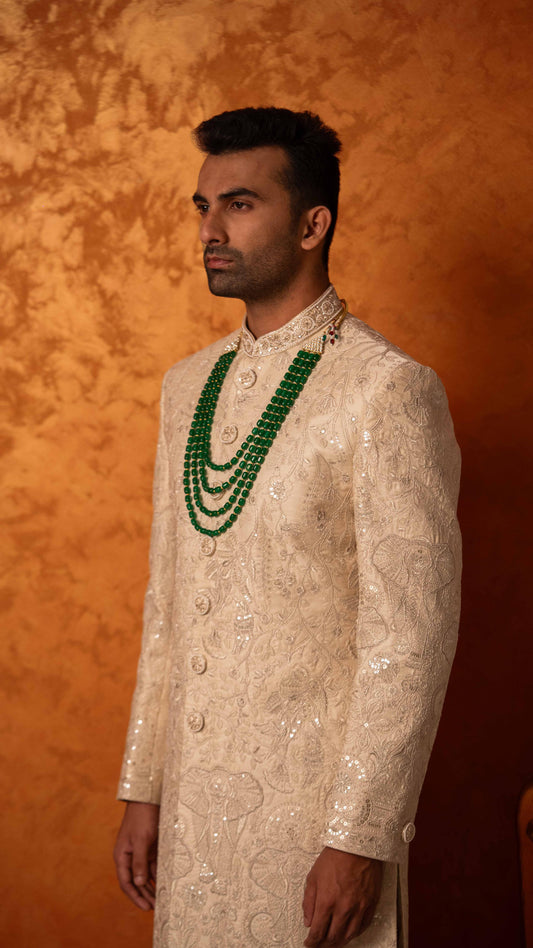 A man wearing a cream-colored embroidered sherwani with a multi-strand green beaded necklace, standing against a textured orange-brown backdrop.