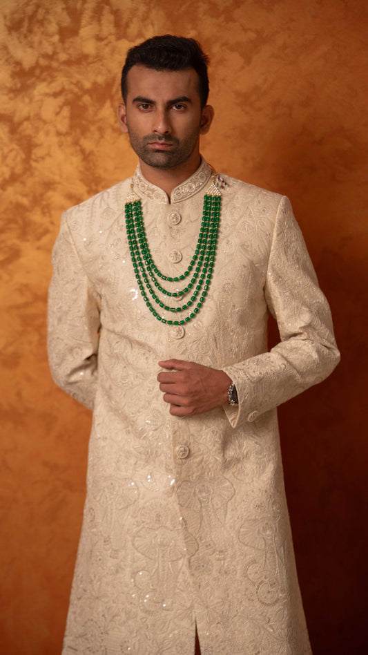 A man wearing a cream-colored embroidered sherwani with a double-row green beaded necklace, standing against a textured orange-brown studio backdrop.