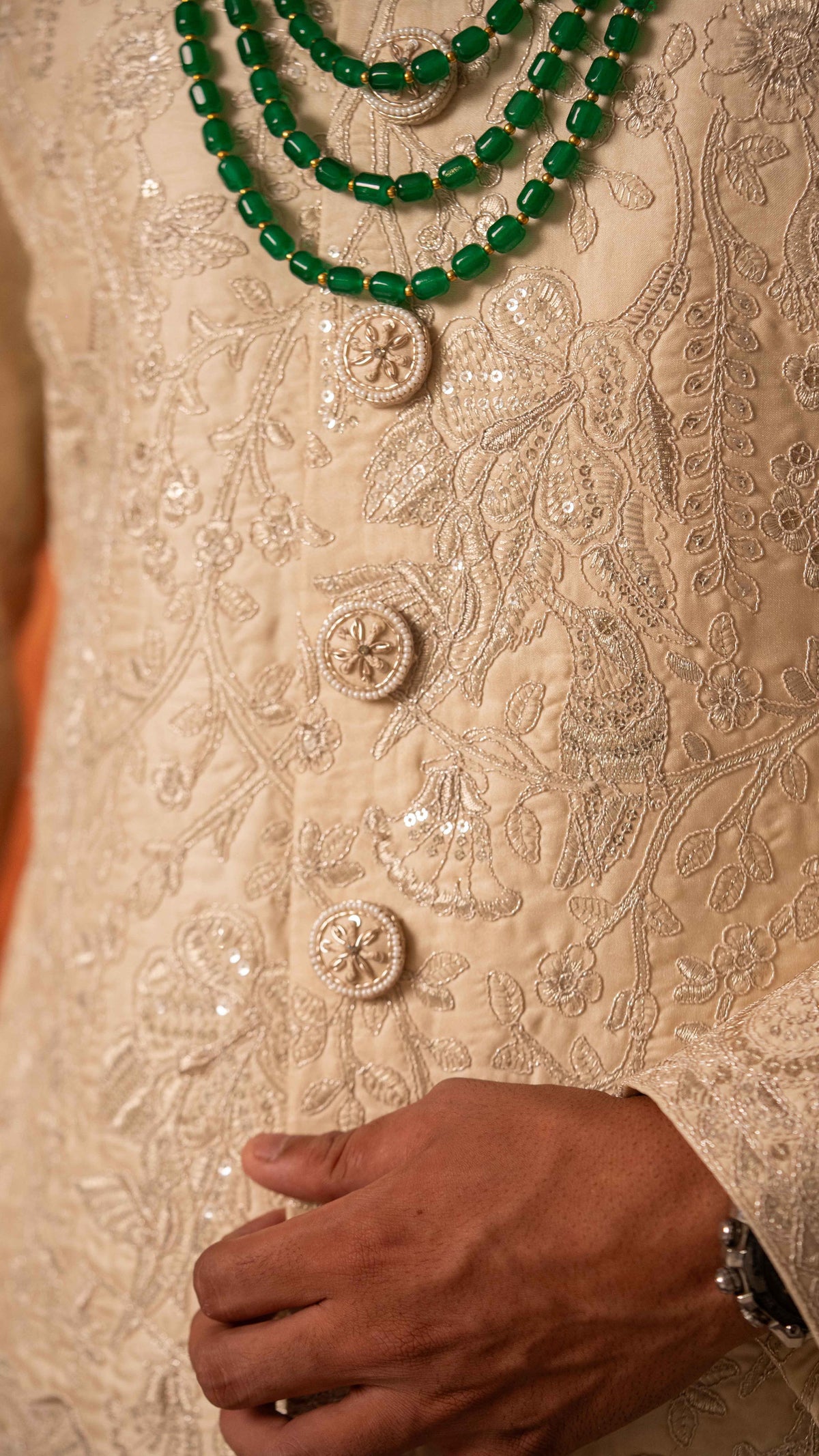 Close-up of a person wearing an ornate cream-colored embroidered traditional garment with sequins and buttons, layered with a green beaded necklace, with a hand resting near the waist.