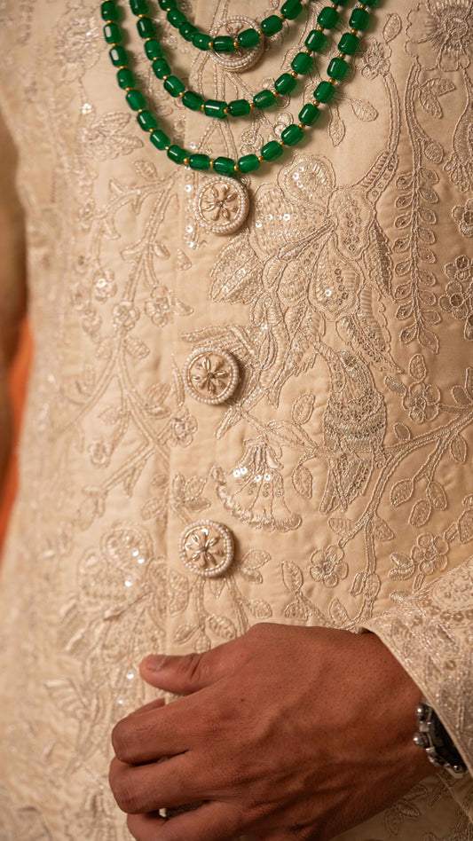 Close-up of a person wearing an ornate cream-colored embroidered traditional garment with sequins and buttons, layered with a green beaded necklace, with a hand resting near the waist.