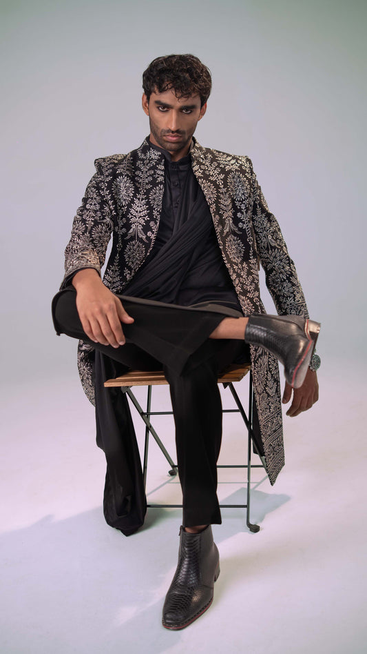 A man sits on a metal folding chair wearing a long black indowestern, a black-and-white ornate patterned jacket, black trousers, and black ankle boots against a light gray studio background.