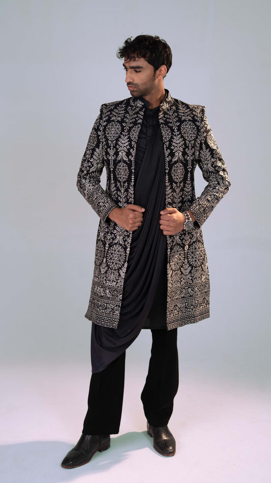 A man modeling a black and grey heavily embroidered sherwani jacket over a black kurta and trousers, standing against a light gray studio background.