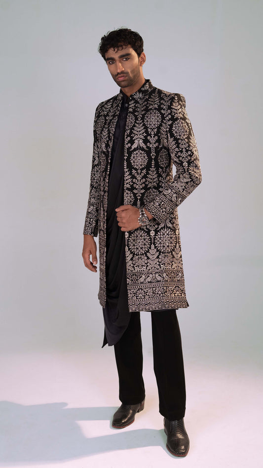 A man modeling a long black and grey patterned sherwani jacket over a black kurta, black trousers, and black dress shoes, standing against a light gray studio background.