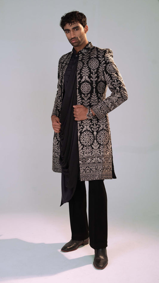 A man modeling a black and grey patterned sherwani jacket over a black kurta, trousers, and black shoes, standing against a light gray studio background.