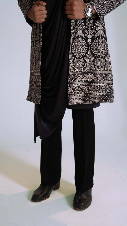 A person wearing a black indowestern with ornate silver and beige floral embroidery, paired with black trousers and black shoes, shown from the chest down against a light studio background.