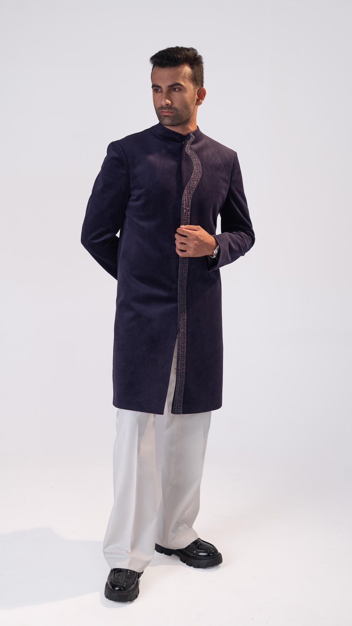 A man modeling a purple indowestern with a decorative metallic border down the front, paired with white trousers and black shoes, against a plain white studio background.