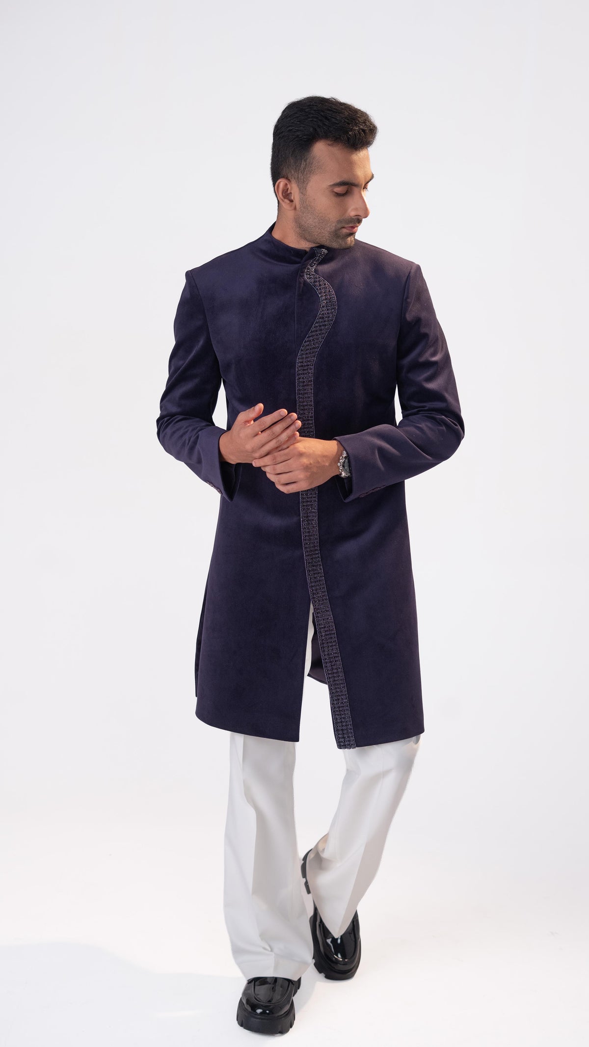 A man modeling a purple velvet sherwani with a textured patterned border, paired with white trousers and black shoes, against a plain white studio background.